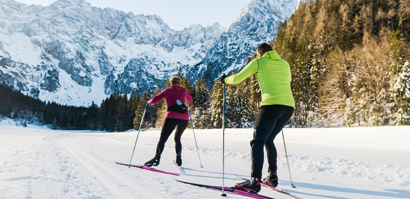 Two cross-country skiers on a groomed trail below snowy peaks