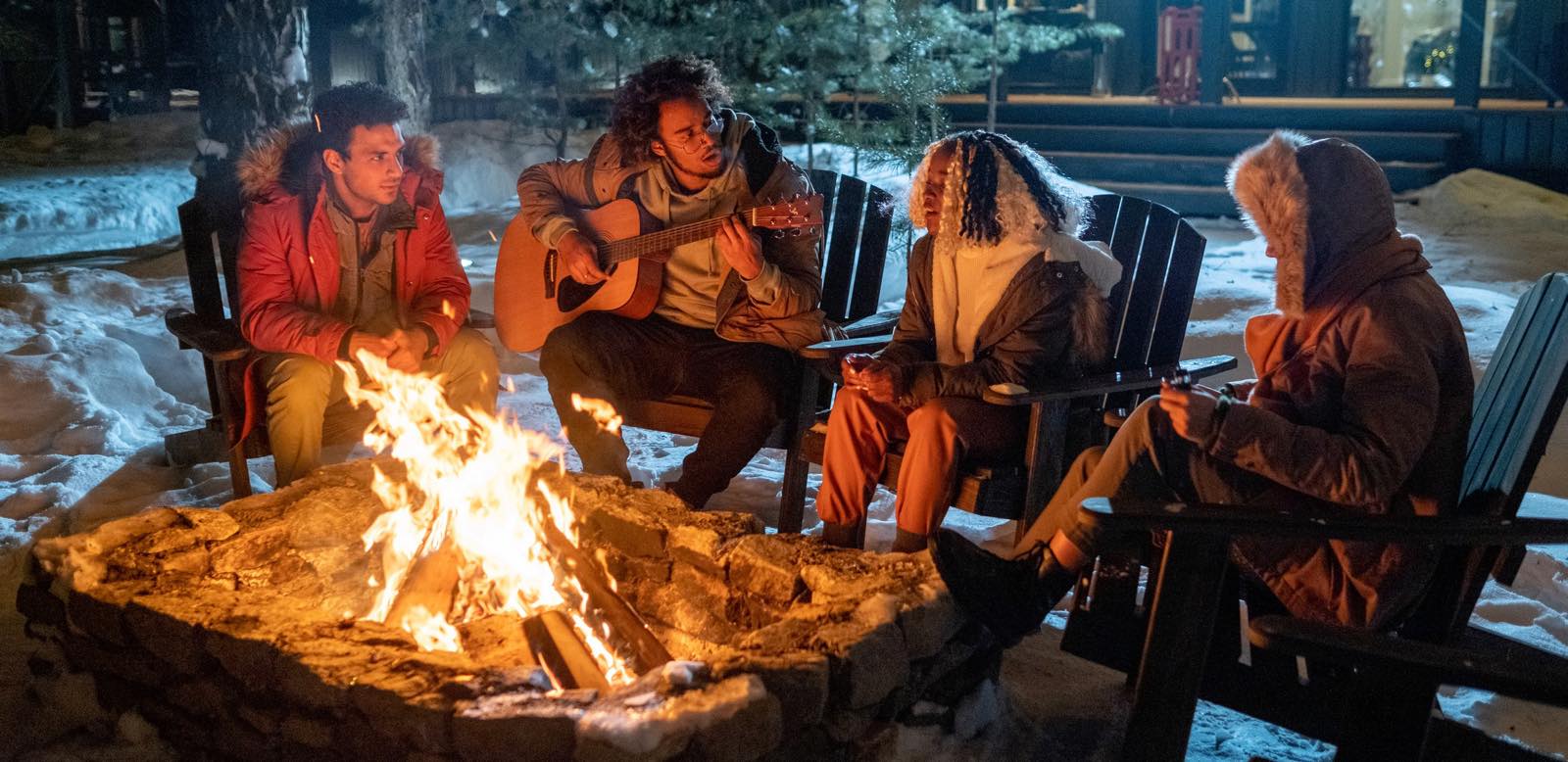 Friends in winter coats gathered around an outdoor fire at night, one playing guitar