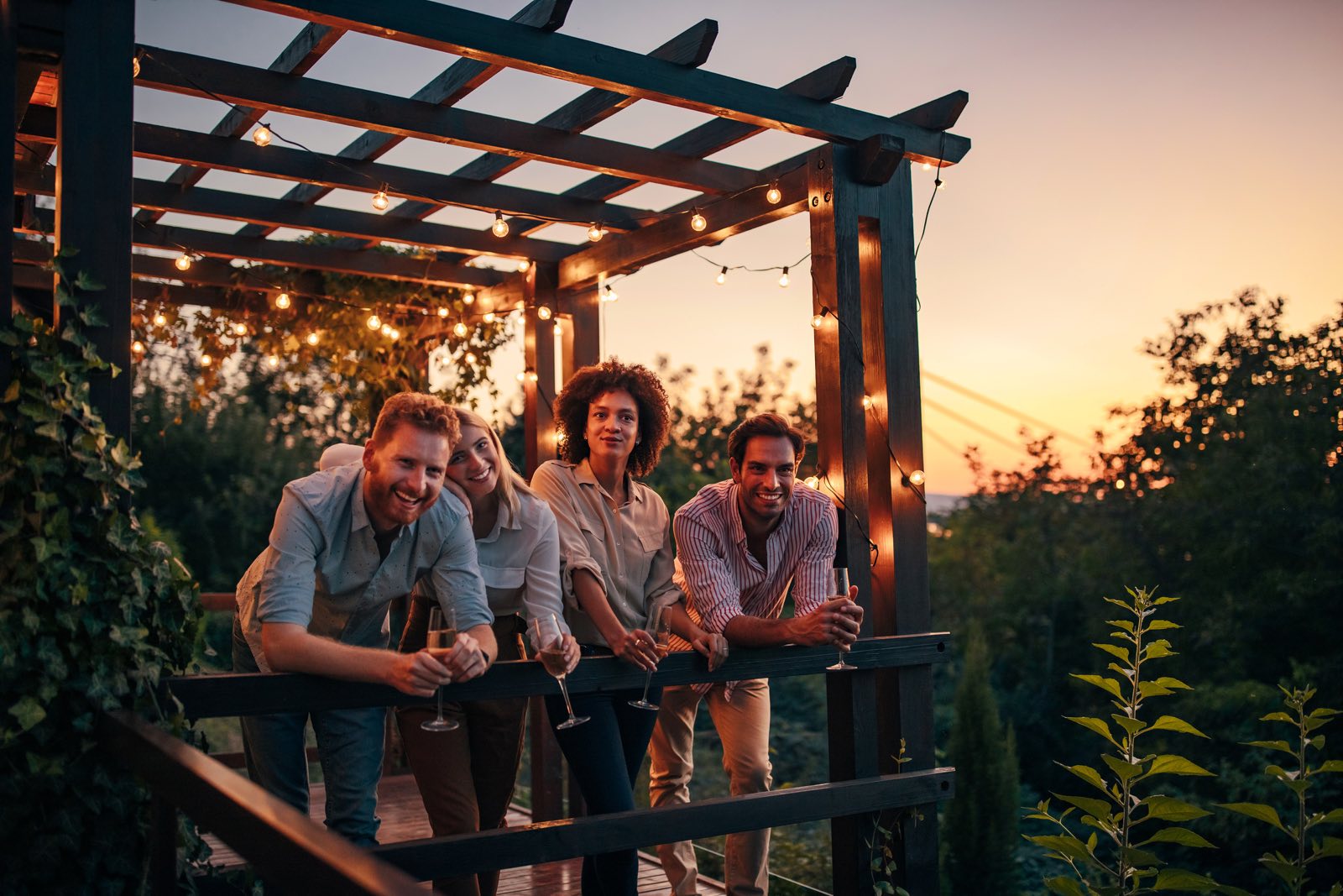 Friends sharing drinks on a deck under string lights at sunset