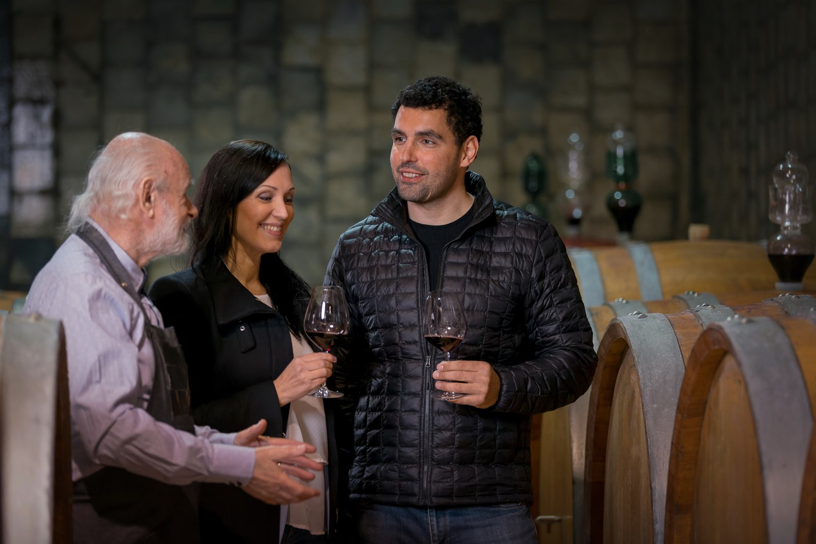 A winemaker pouring for two guests on a cellar tour among oak barrels