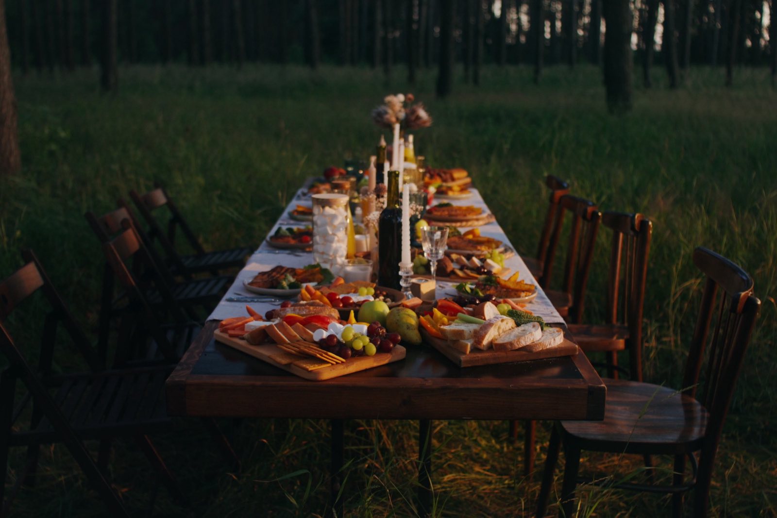 A long candlelit table of food set out in a forest at golden hour